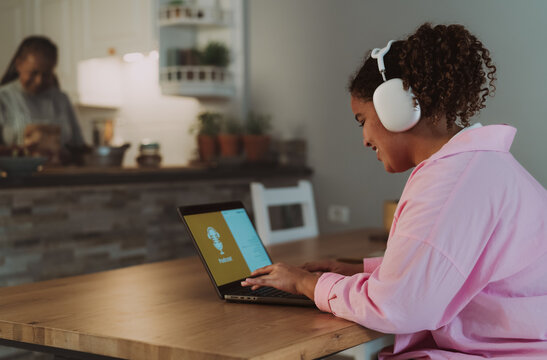 Young African Girl Using Laptop Listening Podcast While Her Mother Cooking In The Background At Home