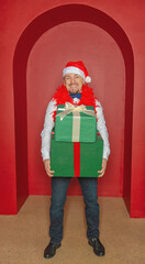 Young handsome smiling man wearing Santa hat holding Christmas gift boxes on the red arch background
