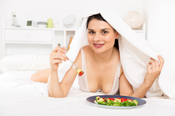 Obraz na płótnie Canvas Positive pretty woman in bathrobe eating vegetable salad under blanket in bed at home ..