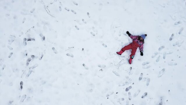 Vista a&eacute;rea desde dron de ni&ntilde;a con ropa de esquiar de colores y gorro de lana lila tumbada en la nieve haciendo el &aacute;ngel 