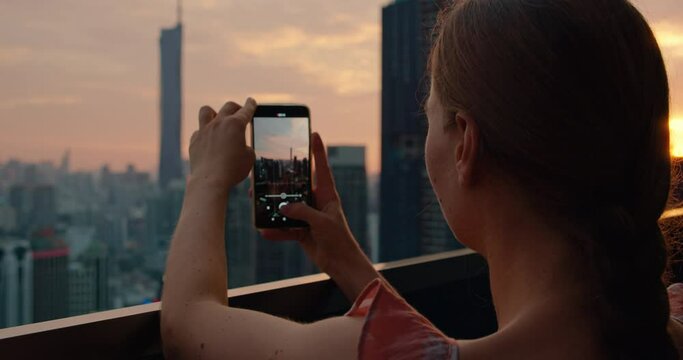 Tourist woman taking photograph of cityscape skyscrapers at sunset in capital of Malaysia. Cloudy twilight peach skyline view from the roof of an apartment building.