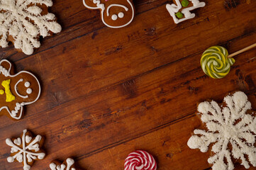 Christmas greeting concept - a brown wooden table background surrounded by various shaped gingerbreads with glaze and caramels with copy space in a middle. Top view.