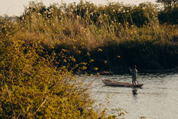 Idyllische Abendszene am Okavango -Ein Junge stakt im Dämmerungslicht mit Einbaum Boot über den...