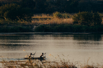Obraz na płótnie Canvas Idyllische Abendszene am Okavango - Zwei Jungen paddeln im Dämmerungslicht mit Einbaum Boot über den Fluss, Rundu, Namibia