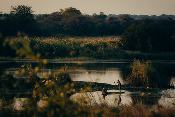 Idyllische Abendszene am Okavango - Zwei Jungen paddeln im Dämmerungslicht mit Einbaum Boot über den Fluss, Rundu, Namibia