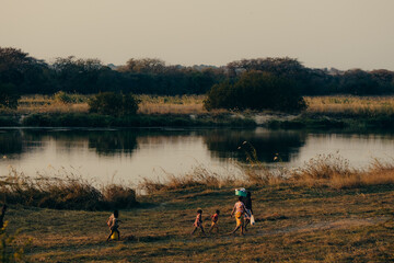 Abendszene am Okavango - Mutter läuft mit ihren Kindern   im Dämmerungslicht, nachdem sie sich am...