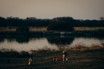 Obraz na płótnie Canvas Abendszene am Okavango - Mutter läuft mit ihren Kindern im Dämmerungslicht, nachdem sie sich am Fluss gewaschen haben, nach Hause, Rundu, Namibia