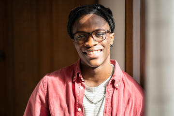 Indoor portrait of happy optimistic young African American guy wearing glasses smiling at camera, standing by window in sunny room at home. Positive attitude and emotional wellbeing concept