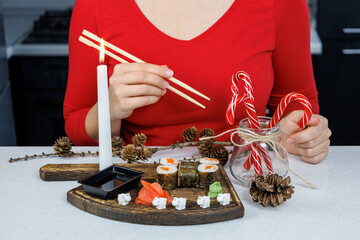 Japanese rolls with red fish on a wooden cutting board. A girl in a red sweater eats sushi with chopsticks. Table decoration with candles and sweets. Christmas concept