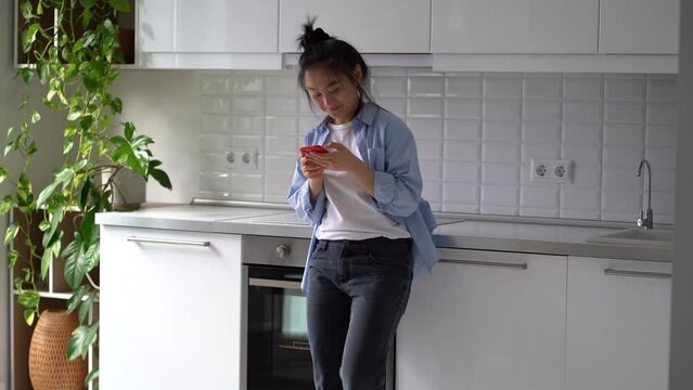 Happy smiling Asian girl standing in kitchen holding smartphone receiving pleasant romantic text message from boyfriend. Young joyful woman chatting in social networks, fallen in love online