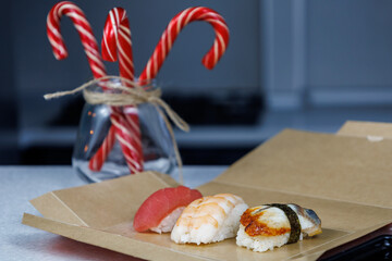 Sushi with tuna, shrimp and eel lies in a cardboard box on the table in the kitchen. Christmas concept. Festive decorations in the form of candles, figurines and sweets