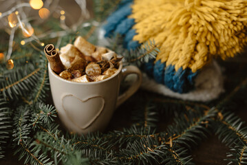 A mug of hot chocolate with cinnamon and marshmallows surrounded by fir branches, garlands and a yellow-blue knitted hat.