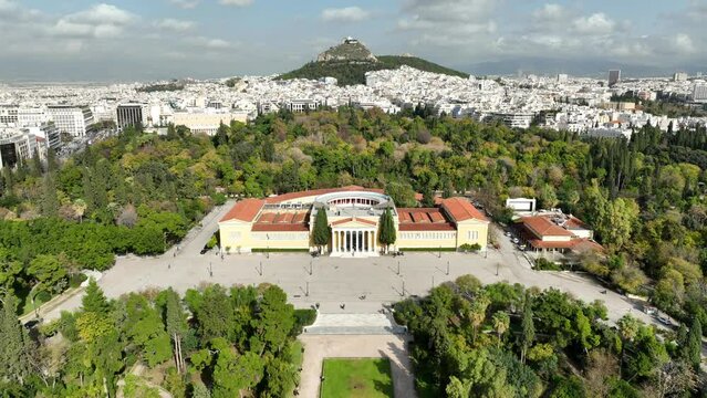 Aerial drone video of renovated public neoclassic building of Zappeion used for events and meetings in the National Gardens of Athens and Lycabettus hill aligned at the background, Greece