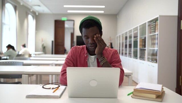 Tired African man sit in library at table with laptop takes off glasses feels pain in eyes stretches and rubs palms. Thoughtful black student at work with computer, thinking about project test exam