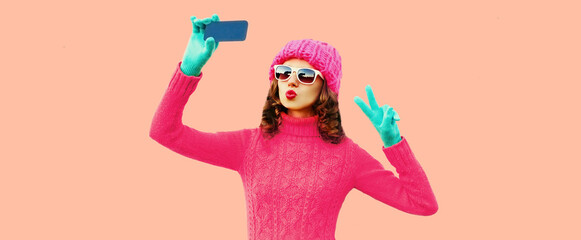 Portrait of stylish young woman taking selfie with smartphone wearing pink knitted hat, sweater on background