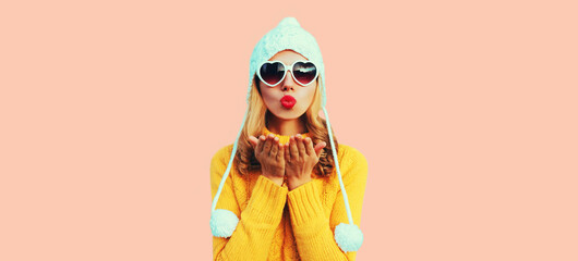 Winter portrait of beautiful woman blowing her red lips sending sweet air kiss wearing heart shaped sunglasses, yellow knitted sweater and white hat with pom pom on pink background