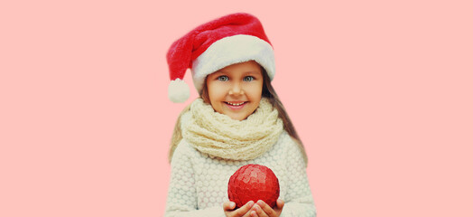 Christmas portrait of happy smiling little girl child in red santa hat with ball toy on pink background
