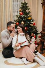 Surprised couple is opening Christmas gift box near the Christmas tree at home at Christmas. 