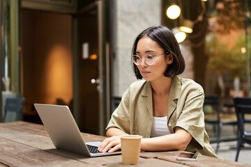 Young woman working in a cafe, using laptop and drinking coffee. Asian girl student with computer studying remotely, sitting on bench near shop