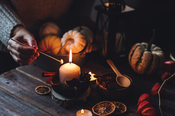 Young woman in the knitted warm sweater lights a candle on a wooden table. Cozy fall atmosphere, beautiful autumn home decor with pumpkins and leaves