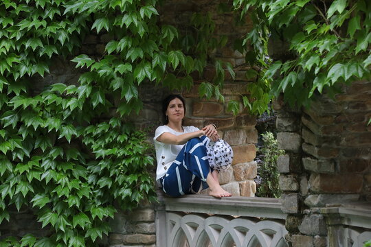 A young woman sitting barefoot on a stone fence leaning against a wall covered with green ivy is resting in the garden in summer