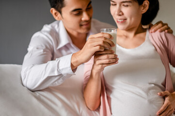 Portrait of Pregnant Asian woman sitting on a white bed Her husband holds a glass of milk and sends her fresh milk in a clear glass for her to drink. To nourish the baby in her womb.
