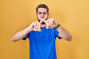 Hispanic man with beard standing over yellow background rejection expression crossing fingers doing negative sign