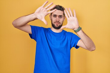 Fototapeta na wymiar Hispanic man with beard standing over yellow background doing frame using hands palms and fingers, camera perspective