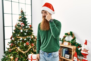 Redhead man with long beard wearing christmas hat by christmas tree pointing unhappy to pimple on forehead, ugly infection of blackhead. acne and skin problem
