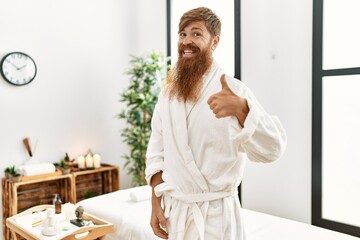 Redhead man with long beard wearing bathrobe at wellness spa smiling happy and positive, thumb up doing excellent and approval sign