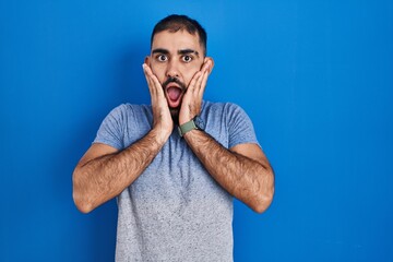 Middle east man with beard standing over blue background afraid and shocked, surprise and amazed expression with hands on face