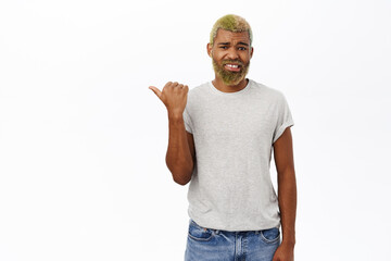 Naklejka premium Dislike. Disappointed african american guy with yellow modern hairstyle and beard, pointing left, grimacing from disappointment, white background