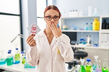 Young caucasian woman working at scientist laboratory holding toxic label covering mouth with hand, shocked and afraid for mistake. surprised expression