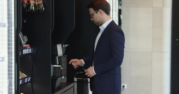 Young businessman in suit prepare coffee, buy snack in vending machine, pay for goods swipe credit card to card reader machine at workday in modern office during lunch break. Services, technologies