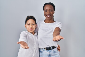 Young mother and son standing together over white background smiling cheerful offering palm hand giving assistance and acceptance.