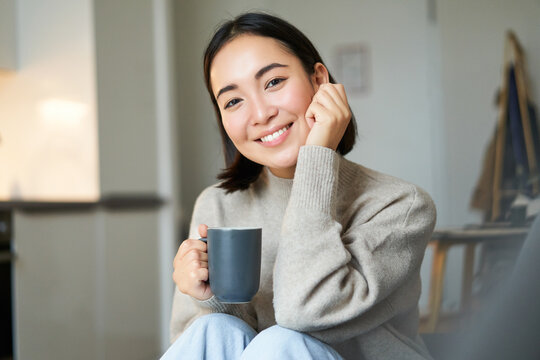 Smiling Asian Woman Sitting At Home With Cup Of Coffee, Relaxing And Feeling Warmth, Looking Outside Window, Resting On Sofa In Living Room