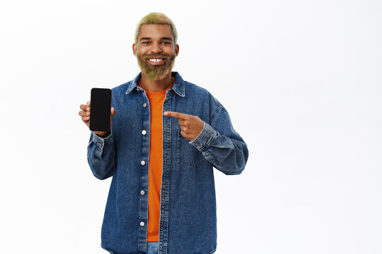 Happy Smiling African American Guy In Stylish Outfit, Pointing At Mobile Phone Screen, Showing Smartphone Display And Smiling Pleased, White Background