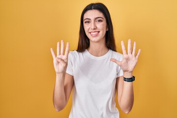 Young beautiful woman standing over yellow background showing and pointing up with fingers number...
