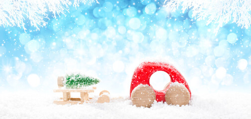 Red Wooden Car Pulling Sled with  Laying Green Christmas Tree in perspective on bright blue bokeh winter background with white snowy branches and falling snow.
