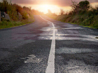 Fototapeta premium Small narrow asphalt road in a country side with slight damage to surface at sunset. Nobody. Selective focus. Travel and tourism concept.