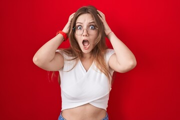 Obraz na płótnie Canvas Young caucasian woman standing over red background crazy and scared with hands on head, afraid and surprised of shock with open mouth