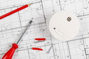 Smoke detector or fire alarm sensor on white architectural plans background with tools and screws, house safety or security concept, flat lay top view