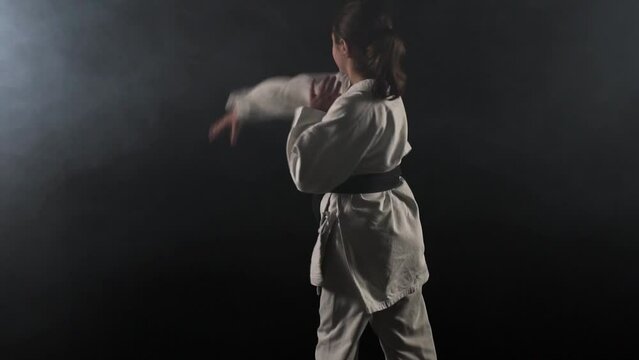 Young karate girl wearing kimono posing and spinning against black background with smoke. Slow motion recorded at 100fps