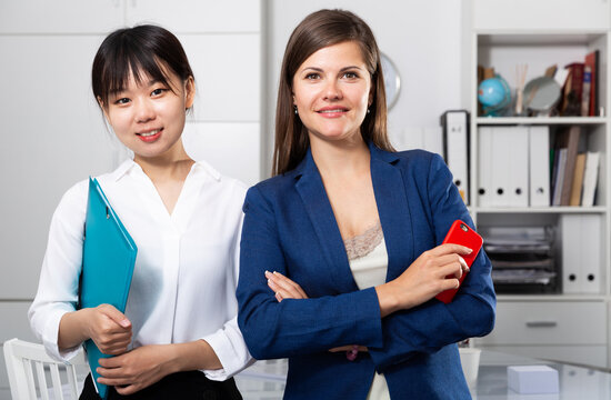 Two confident women members of multinational team posing in office of international company