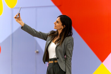 Glad adult businesswoman in smart casual clothes with dark hair smiling and taking selfie against...
