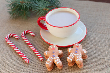 Hot winter drink - chocolate in white mug with gingerbread men and candy canes. Christmas time. Cozy home atmosphere, burlap fabric background. Holiday mood in the air