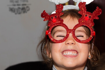 Little brown-haired Caucasian girl wearing red Christmas-style glasses. Little girl with funny glasses. Christmas complement
