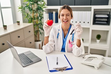 Young blonde woman wearing doctor uniform holding blood and heart winking looking at the camera...