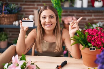 Young blonde woman working at florist shop holding credit card smiling happy pointing with hand and finger to the side