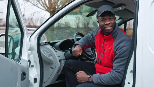 Young adult black delivery man wearing red pullover and black cap sitting in door of white van smiling showing thumbs up to the camera. High quality 4k footage